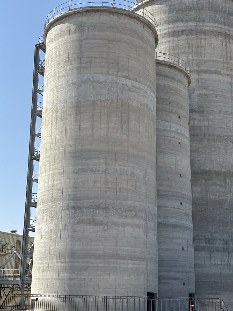 Tall concrete cement storage silos with an external access stair tower, seen from ground level at a cement plant